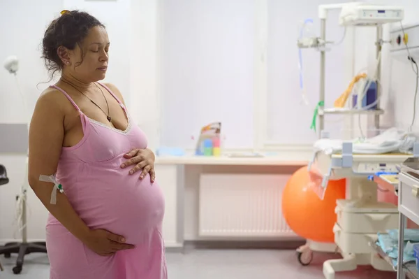Multi-ethnic birthing mother having contraction, holding her big belly in the labor room of maternity hospital. Pregnancy. Easy delivery. Obstetrics and gynecology. Childbearing and childbirth concept