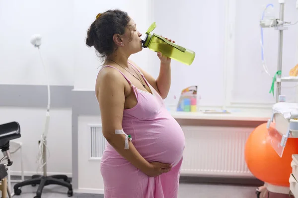 Side view to a pregnant woman birthing mother with painful contraction, touching belly and drinking water in a hospital ward. Childbirth process