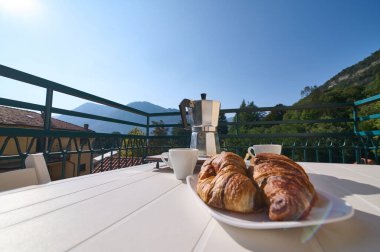 Taze kruvasan tabağı, İtalyan Alpleri 'nin arka planında taze demlenmiş kahve makinesinin yanında beyaz fincanlar. İtalyan yaşam tarzı ve kültürü. Kahvaltıda. Günaydın konsept..