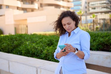 Çekici Latin kadın, menajer, girişimci, mavi gömlekli, internette sörf yapan, akıllı telefonundaki mobil uygulamaları kontrol eden, web sitelerini gezen, modern binalarla iletişim kuran,