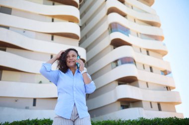 Çok ırklı güzel bir kadın, girişimci, 30 'lu yaşlarda mavi gömlekli bir satış müdürü, cep telefonuyla konuşurken gülümsüyor, modern binaların arka planına karşı. İş için. - İnsanlar. İletişim