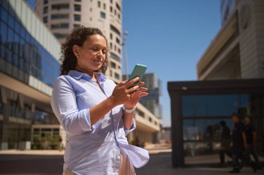 Modern akıllı telefonunu elinde tutan bir kadın girişimcinin alt görünümü, yeni mobil uygulamaları kontrol etmek, rezervasyon, sipariş vermek, web sitelerini taramak, online rezervasyon yapmak, açık havada durmak,