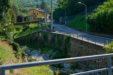 Canzo, Lombardy, İtalya. Eylül 2023. Seyahat ve turizm konsepti. Dağlardaki sarı renkli güzel evin manzarası ve İtalya Alpleri 'ndeki bir dağ nehrinin üzerinden geçen yol.