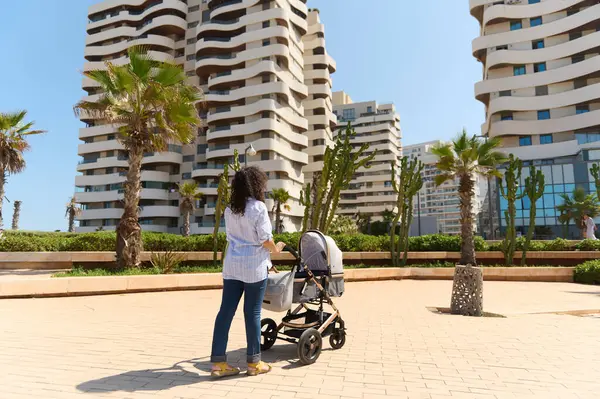 Back view of a curly haired woman with her baby in pram, strolling the street in an urban city with modern high rise buildings on the background. Maternity leave and babyhood concept