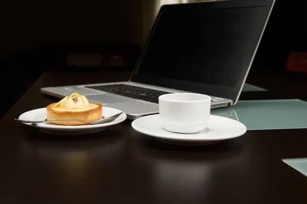 Still life with food and digital gadget. A sweet delicious tartlet with lemon custard, a cup of freshly brewed espresso coffee over the background of a laptop with black blank monitor screen.