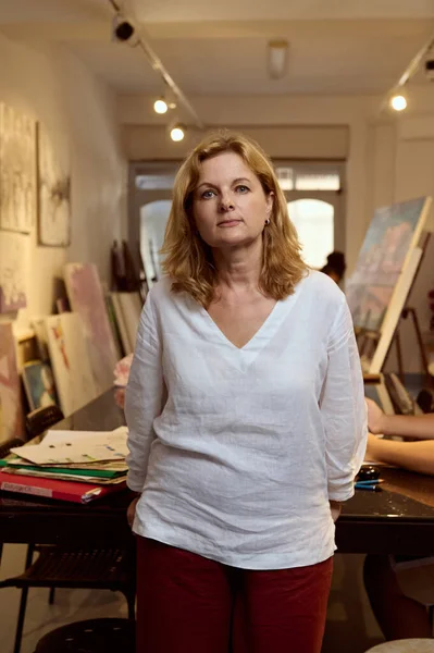 Professional portrait of beautiful mature woman, artist, painter, teacher in the art workshop, looking at camera, standing in her creative art studio with displayed painted pictures and reproductions