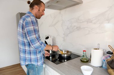 Gayri resmi giyinmiş yakışıklı beyaz bir adam mutfak tezgahının yanında duruyor ve ev mutfağında yemek hazırlarken malzemeleri kızartma tavasına karıştırıyor. İnsanlar ve boş zaman aktiviteleri