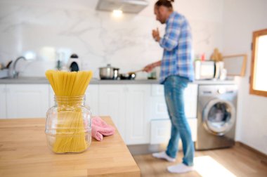 İtalyan makarnasına odaklan. Evde yemek pişirirken mutfak tezgahında duran yakışıklı bir adamın arka planına karşı şeffaf cam bir kabın içindeki spagetti. İtalyan mutfağı