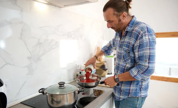 Attractive handsome chef hand holding a bottle with passata and adding tomato sauce into a frying pan with olive oil and bay leaf, cooking pasta for dinner, according Italian traditional family recipe