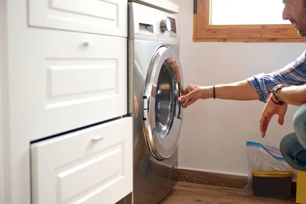 Handsome Caucasian bearded guy dressed casual, crouching in home bathroom and loading washing machine with dirty clothes. It\'s laundry day. Man loading washing machine. Housekeeping. Household. Chores