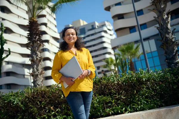 Beautiful confident businesswoman in casual clothes, holding a laptop, smiling looking at camera, standing against high-rise modern buildings background. People. Work. Business. Freelance occupation