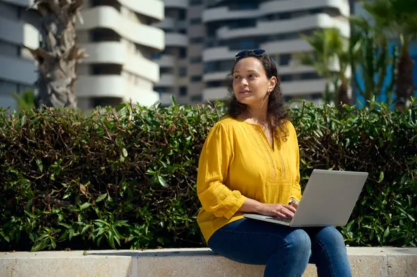 Dizüstü bilgisayar kullanan, online çalışan, şirket kurmayı planlayan, gülümseyen, taştan bir bankta modern kentsel binalara karşı otururken kenara bakan serbest çalışan bir kadın girişimcinin özgüvenli portresi.