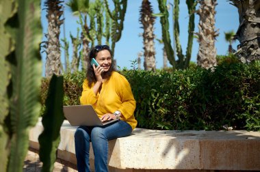 Dizüstü bilgisayarlı çok ırklı, kendinden emin genç bir iş kadını, doğa parkında taş bir bankta oturuyor, iş ortaklarıyla cep telefonunda konuşurken yan yana bakıp gülümsüyor. - İnsanlar. İletişim.