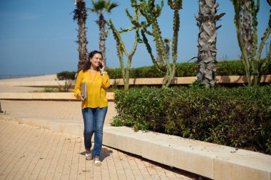 Kendine güvenen çok ırklı iş kadını, gündelik giysiler içinde, akıllı cep telefonuyla konuşurken geniş bir şekilde gülümsüyor ve elinde bir dizüstü bilgisayarla sokakta dolaşıyor. Tam boy portre. İş Kariyeri
