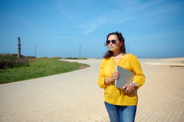 Güzel Latin Amerikalı kıvırcık esmer, güneş gözlüğü takan serbest çalışan, sokakta dolaşan, elinde dizüstü bilgisayarla ofise giden bir kadın. - İnsanlar. Yaşam tarzı. İş için. İşe alım ve kariyer.