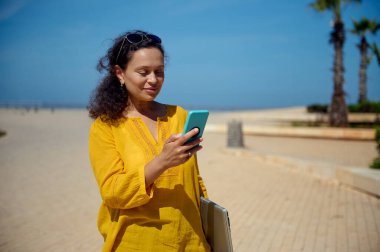 Gülümseyen, çoklu etnik çeşitlilikte mutlu bir kadın akıllı telefondan cep telefonu uygulamasını kontrol ediyor, dizüstü bilgisayarını elinde tutuyor, gezinti güvertesinde geziniyor, ofise gidiyor. Cep telefonu kullanan güzel bir esmer.