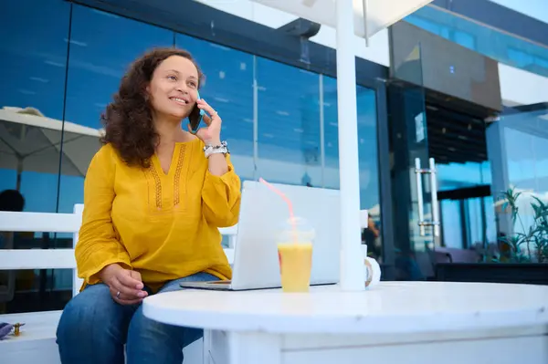 Çok ırklı, orta yaşlı, güzel bir kadın açık dizüstü bilgisayarlı bir kafede oturuyor, cep telefonuyla konuşurken gülümsüyor. - İnsanlar. Çevrimiçi iş ve uzaktan çalışma kavramı