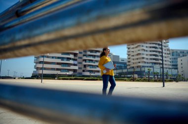 Kendine güvenen genç bir iş kadını, sıradan giyinmiş girişimci, dizüstü bilgisayarı olan ve cadde boyunca yüksek binaların arkasında yürürken yan tarafa bakan biri. Şehir yaşam tarzı. - İnsanlar. Kariyer