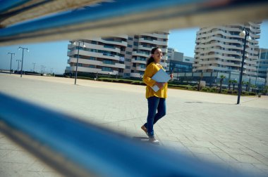 Gündelik giysiler içinde, elinde dizüstü bilgisayarı olan ve kameraya bakıp gülümseyen, modern gökdelenlerin arka planında gezinen kendine güvenen genç bayan girişimci. - İnsanlar. İş için. Yaşam biçimi