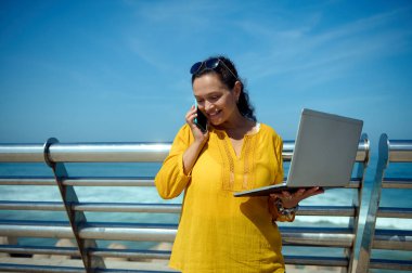 Gülümseyen güzel bir girişimci kadın cep telefonuyla konuşuyor, iş ortaklarıyla görüşüyor, dizüstü bilgisayarı tutuyor ve dışarıda çalışıyor. Serbest meslek işte. Uzaklık çalışması