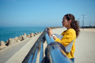 Akıllı telefon kamerası kullanan, deniz manzarasını fotoğraflayan, gezinti güvertesinde dizüstü bilgisayarla duran çok ırklı, çok ırklı genç bir kadın.