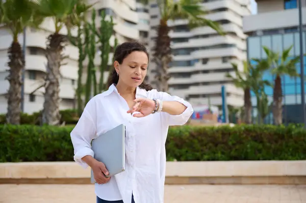 Authentic waist-up portrait of a woman entrepreneur checking time on her wrist watch, waiting for important business deal, holding a laptop,standing outdoor against modern high rise buildings backdrop