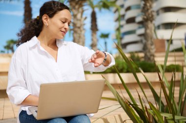 Beyaz günlük gömlekli başarılı bir iş kadını, kol saatine bakıyor, dizüstü bilgisayarla yüksek binalarda çalışıyor. - İnsanlar. Emlak yatırımı. Uzak görev