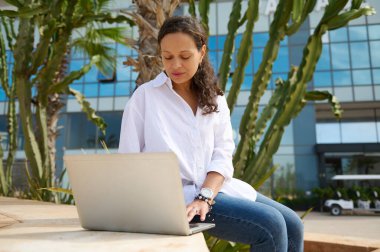 Mavi kot ve beyaz gömlekli Latin Amerikalı genç kız öğrenci, dizüstü bilgisayarda yazı yazıyor, online olarak diploma projesi üzerinde çalışıyor, merdivenlerde oturuyor, modern yüksek binaların arka planına karşı.