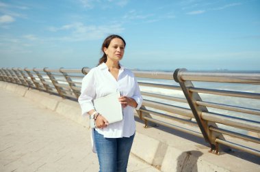 Gündelik giysiler içinde genç düşünceli Latin bir kadın dizüstü bilgisayarını tutuyor ve deniz kenarında yürürken dikkatlice uzaklığa bakıyor. - İnsanlar. Uzaktan kumanda. Çevrim içi iş