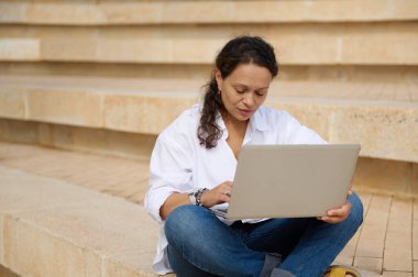 Çok ırklı esmerin kendine güvenen portresi, dışarıda merdivenlerde oturan genç yetişkin bir kız öğrenci, dizüstü bilgisayarla yeni diploma projesi üzerinde çalışıyor. - İnsanlar. Yaşam tarzı. Kariyer. Çevrimiçi Eğitim. Üniversite