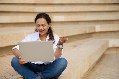 Gülümseyen esmer, beyaz gömlekli ve kot pantolonlu Latin Amerikalı güzel bir kadın laptopla video görüşmesi yapıyor, dışarıda mermer merdivenlerde otururken el kol hareketi yapıyor. - İnternet. Çevrimiçi iletişim