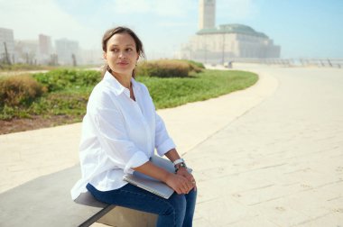 Çekici genç bir kadın dizüstü bilgisayarla bankta oturuyor, öğle arasında dinleniyor. Düşünceli iş kadını, mesafeye dikkatlice bakıyor, beyaz günlük gömlek ve kot pantolon giyiyor.