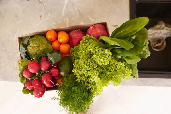 View from above of a recyclable cardboard box with fresh organic crop of fruits, vegetables and greens: broccoli, spinach, radish, dill, tomatoes, tangerines and pomegranates on the kitchen counter