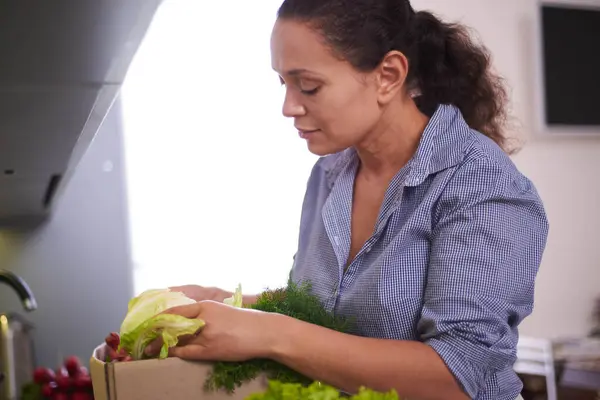 Online home food delivery. Multi ethnic woman checking her online order list. Cardboard box with fresh vegetables and fruits standing on the kitchen table. Local farmer food. Start of a healthy life