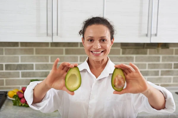 Multi ethnic smiling woman looking at camera and showing two halves of fresh ripe organic avocado fruit, standing against gray brick wall background in the home kitchen. Alimentation and healthy food