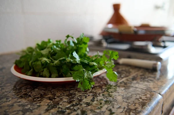 Closeup of coriander and parsley on a plate on marble kitchen counter, against the background of a tagine clay pot cooking on stove in the kitchen at home. Herbs and greens. Healthy food concept