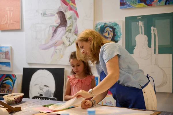 Caucasian charming young woman teacher wearing blue apron, standing by desk, explaining drawing technique to a little girl student while teaching art at creative studio. Kids. Creative art education