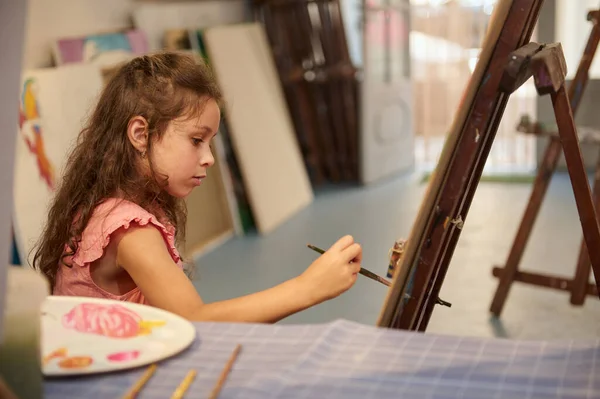 Adorable inspired Caucasian child, little girl artist painting on canvas doing some art projects on a creative studio workshop. Elementary age child learning painting in modern fine art studio