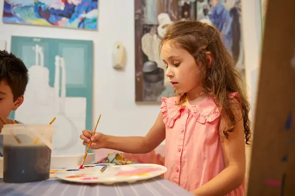 Caucasian adorable kid girl learns drawing, standing by desk with palette of watercolors, against a wall with exhibited pictures in fine art gallery or creative workshop. Painting. Education. Children