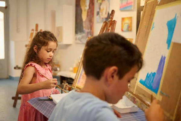 Creativity, inspiration, art and painting concept. Adorable talented kids, teenage boy and schoolgirl, little artists standing in front of easel, holding brush and palette, drawing in fine art studio