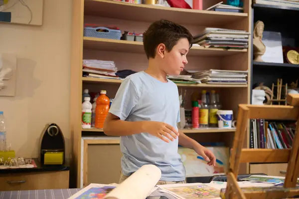 Smart student boy learning drawing in creative art studio. Handsome confident little artist painter standing by desk near a wooden easel, choosing sketch for making image reproduction with watercolors