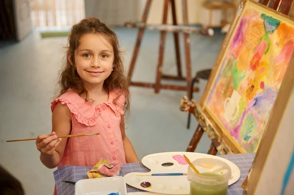 可爱的小女孩艺术家带着可爱的微笑看着摄像机 坐在室内图片旁边 同时在一个创意工作室学习美术 孩子们教育 学习绘画和绘画 — 图库照片#