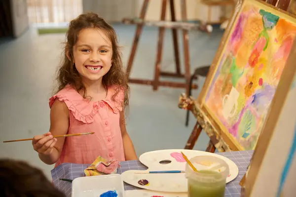 カカシアンかわいい小学生の子供 小さな女の子アーティストは 美しい歯並みのある笑顔で微笑み カメラを見て 絵筆を持ち クリエイティブスタジオで美術を学んでいる間にキャンバスに描きます — ストック写真