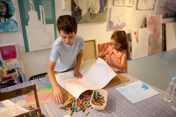Overhead view kids drawing in fine art studio. Adorable teenage boy and little girl using colorful pencils, making a colorful background on white cardboard paper sheet. Visual art. Hobbies and leisure