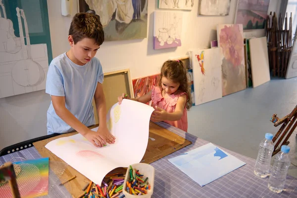Authentic portrait of two adorable kids, a preteen boy and little girl, brother and sister drawing, enjoying creative hobby during an art class in a workshop. Children, creativity and fine art concept