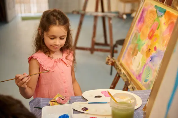 Creativity, inspiration, art and painting concept. Portrait of positive talented little child girl artist wearing apron, standing in front of easel, holding brush and palette, working in fine art studio