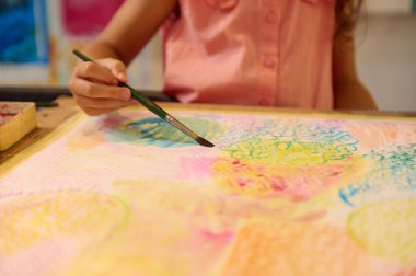 Küçük kız ressamın eli masa başında elinde boya aksesuarları, elinde fırça ve çizim ile oturuyor. Suluboya atölyesi. Sanatsal Uyanış. Çocuk suluboya resmin güzelliğini kucaklıyor