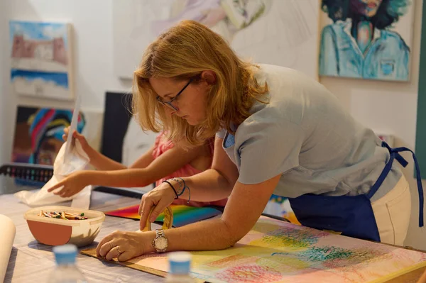 Inspired creative middle aged woman artist, art teacher during her work in art gallery or workshop. Female painter teaching a little student, developing imagination and creativity in fine art studio