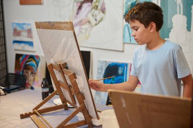 Yakışıklı, çekici, ilkokul çocuğu mavi günlük tişörtlü, tahta sehpası olan, elinde boya fırçası tutan ve sanat dersinde akrilik boyalarla resim yapan.