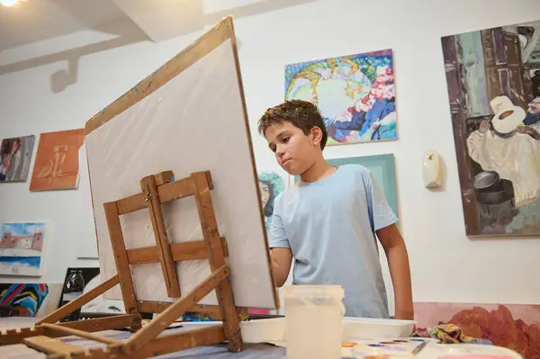 Handsome Caucasian teenage boy drawing on canvas, standing by wooden easel at art class, against the background of painted pictures exhibited on a white wall in the art gallery of visual art school